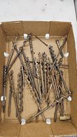 Box of mixed large metal drill bits with differing thicknesses and lengths, some showing rust.