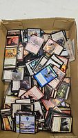 Photo of a cardboard box filled with a large assortment of Magic the Gathering cards loosely sorted and stacked, showing many different card backs and fronts.