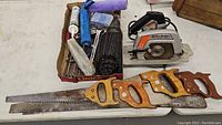 Mixed tool lot showing circular saw, handsaws, and box of miscellaneous items on white floor