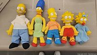 Full view showing all five Simpsons dolls: Homer, Marge, Bart, Lisa, Maggie, arranged side by side holding hands.