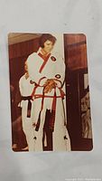 Full view of a color photograph showing Elvis Presley wearing a white karate gi with red trim and black belt with red markings, standing indoors with wood-paneled background, hands clasped.