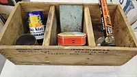 Wooden box divided into compartments containing various vintage tins: Sunkist lemonade tin, Players tobacco tin, a large cylindrical fly spray can, and small metal tins.