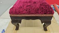 Top corner close-up of footstool upholstery and leg detail showing burgundy fabric and carved wooden leg with scalloped apron.