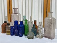 Full collection arrangement of 15 antique bottles in varied colors including cobalt blue, green, brown, light purple, and clear glass on white surface.