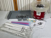 Full view of Nostalgia cotton candy maker with accessories arranged on a white table including scoops, packaged white paper cones, plastic cones, instruction manual, and packaging tray.