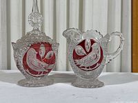Covered sugar bowl with lid and cream pitcher, ruby red and clear glass with bird motifs, set on white background
