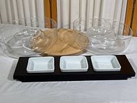 Two large clear glass chip and dip bowls, a round wood serving platter, and an 18-inch wooden tray with three white rectangular condiment dishes arranged on a white surface.