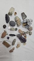 Overview of multiple natural rocks, crystals, and minerals, including amethyst cluster and geode pieces, laid out on white fabric.