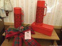 Two red and black plaid Aladdin thermoses with matching solid red lunch box on wooden surface against wallpaper background.