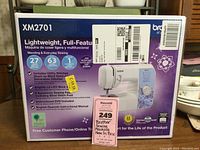 Box front showing Brother XM2701 sewing machine model, features, and sticker tags including Maxsold price tag.