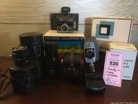 Group photo of vintage cameras and lenses showing Kodak Six-20 Bullseye, Polaroid Colorpack II box, two Canon FD lenses in cases, and other camera accessories.