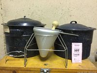 Two black speckled canning kettles with lids, metal perforated ricer with wooden handle on stand, metal jar lifter in front.