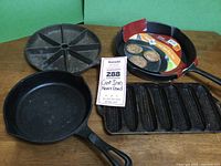 Overview of four cast iron cookware pieces on a wooden table, including skillet, corn grill pan, divided wedge pan, and new fry pan with packaging.