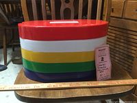 Side view of four stacked plastic picnic trays in red, yellow, green and blue with a red snap-on handle cover, placed on a ruler for size reference.
