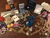 Wide view showing majority of costume jewelry lot spread on a table including pearl bracelets, colorful beaded bracelets, earrings in boxes, gold-tone pendants and chains.
