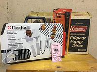 Three items displayed on a table: Char-Broil grilling tool set in closed white case, Thermo Fork in original packaged blister pack, Coleman Deluxe two burner propane stove boxed.