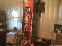 Tall boxed Christmas decoration with Mickey Mouse and friends on front and side of box in room with vintage clock and window.