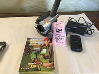 Sony Handycam TRV 130 camcorder with accessories including rechargeable battery in bag, remote control, power adapter, and user manual on display surface.