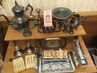 Photo of all items on two levels of wooden shelves showing the candle holders , teapot, silver trays, salt and pepper shakers, and boxed silver knife handles and utensils sets