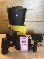 Photo displaying both pairs of Tasco binoculars along with their cases and box. Large binoculars positioned front left with leather case behind and yellow Tasco box beneath. Small binoculars sit right next to large pair with small black fabric case behind.
