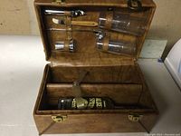 Open leather carrying case showing two plastic glasses, metal bar tools, and padded sections for liquor bottles inside.