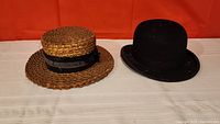 Both antique hats side by side on a white cloth with orange background, showing the woven straw hat with ribbon and the black bowler hat.