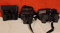 Three sets of birdwatching binoculars with cases on a table, black color, textured grip, and straps visible.
