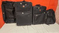 Four black Swiss Gear luggage pieces: large rolling suitcase, two smaller rolling suitcases, and a backpack lined up side by side on a white surface with orange background.