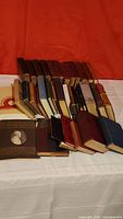 Overview of box lot containing about 22 old books with varying sizes, colors, bindings, some leather bound, arranged on surface with red background.