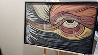 Framed artwork leaning on easel showing entire piece front view. Depicts abstract eye with ribbon-like strokes in pastel.