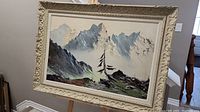 Full view of the Otto Rut oil painting depicting a mountain landscape with the Three Sisters peaks, showing the ornate carved cream-colored wooden frame around the artwork.