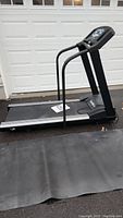 Full side view of black Pacemaster treadmill with handrails and large running belt placed on black rubber mat outside garage door.