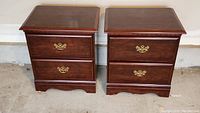 Pair of two drawer wooden nightstands with brass handles, dark brown finish, flat tops, and scalloped bottom edges.