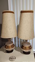 Pair of retro Mid-Century Modern style table lamps with beige fabric shades and decorated ceramic bases on round teak bases.