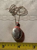 Photo of the oval sterling silver locket pendant on chain showing floral engraving and partial measuring tape for scale.