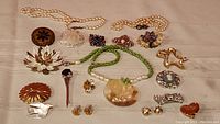 Overview of the lot showing pearl necklaces, various brooches, earrings, and green beaded necklace laid out on a beige surface.