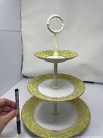 Front view of the dismantlable 3 tier porcelain cake plate with gold rims and green floral pattern, showing the three plates stacked on a cream-colored stand with a handle on top. A pen is held nearby for scale.