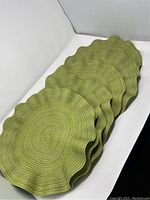 Stack of eight round woven green and gold placemats with scalloped edges showing the stacked profile and texture of the woven material.