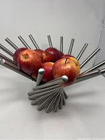 Front angled view of the stainless steel fruit bowl holding several apples, showing overall structure and spoke ends, one missing cap