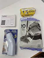 Four items on a white surface: Reynolds Handy Vac sealed in plastic packaging, Shark Plus vacuum bags in a purple and white pack, Yukihalu ceiling fan remote control kit box, yellow Magic flame packs.