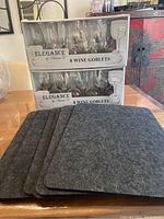 Boxes of 16 unopened Elegance by Vinum 30 wine goblets stacked on top of 6 charcoal gray felt placemats on a wooden table.