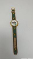 Full length view of Hanslin watch laid flat, showing green leather embroidered band with yellow horse and jockey images and gold-tone watch face showing horse racing design.