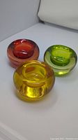 Three round colored glass tealight candle holders in red, yellow, and green with glossy finish, placed on a white surface.
