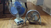 Wide shot showing both the taller blue-bladed Torcan fan on the left and the smaller metal vintage style fan on the right on a wood floor in a room.