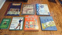 Seven books laid out on wooden floor: 'The Timechart History of Jewish Civilization,' 'Writers,' 'A Day in the Life of the Maple Leafs,' two golf humor books, Home Improvement guide, and Bible stories.