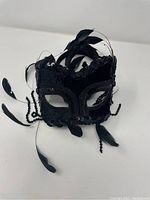 Front angled view of the black velvet masquerade mask with feather and sequin decorations, showing the headband for attachment.