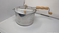 Side view of the popcorn maker showing aluminum kettle and attached wooden hand-crank handle.