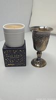 Silver plated wine goblet with grapevine relief decoration next to new Nespresso Pierre Hermé scented candle in a white ceramic container on its black and gold box.