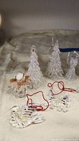 Photo shows 10 clear glass Christmas ornaments including 2 Christmas trees, 2 angels, bell (broken), dove, star, snowflake, candy cane, wrapped gift, laid out on light fabric.