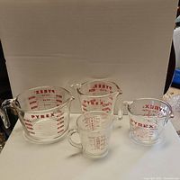 Set of four clear glass Pyrex measuring cups with red markings in various sizes on a white background.